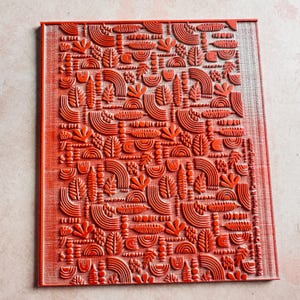 May include: A rectangular, red rubber stamp with a repeating pattern of stylized leaves, arches, and other organic shapes. The design is raised, creating a textured surface. The stamp is likely used for crafting or art projects.