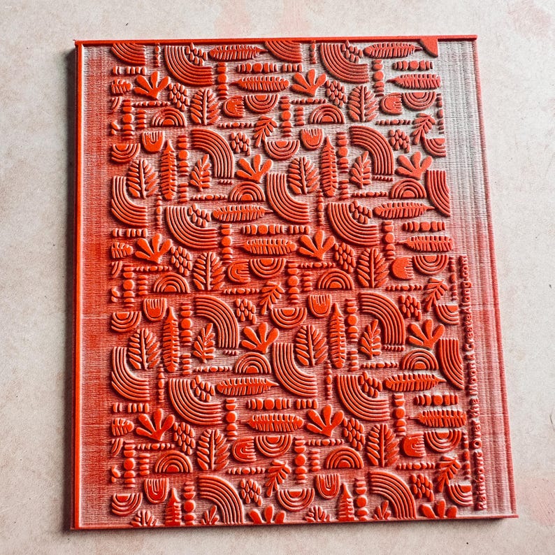 May include: A rectangular, red rubber stamp with a repeating pattern of stylized leaves, arches, and other organic shapes. The design is raised, creating a textured surface. The stamp is likely used for crafting or art projects.