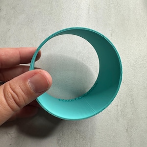 May include: A circular teal plastic cookie cutter with a smooth, flat edge. The side of the cutter features embossed text that reads "Team Perfect Cut" in a sans-serif font. The object is a single, continuous ring shape designed for baking.