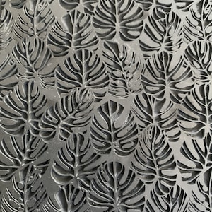 May include: A close-up of a black and white patterned surface with a repeating design of monstera leaves. The surface is textured and has a slightly metallic sheen.