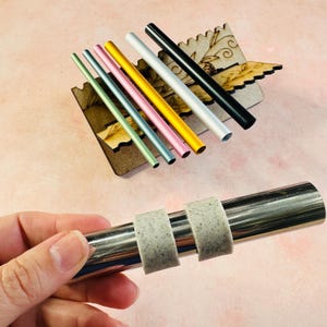 Pode incluir: Um conjunto de oito canudos de metal coloridos em um suporte de madeira, com um close-up de um canudo de aço inoxidável com duas pontas de silicone.
