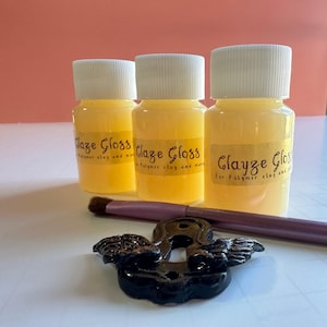 Puede incluir: Tres frascos pequeños de barniz brillante transparente amarillo para arcilla polimérica. Los frascos tienen tapas blancas y etiquetas amarillas con el texto "Clayze Gloss" y "Para arcilla polimérica y más".
