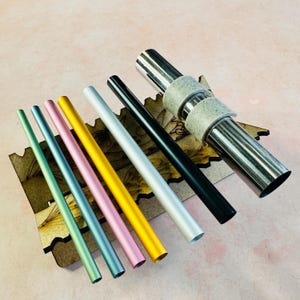 May include: Assortment of eight metal drinking straws in various colors, including green, blue, pink, gold, silver, and black. The straws are arranged on a wooden stand with a silver metal straw holder.