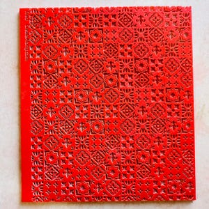 May include: A bright red, square craft stencil with a repeating pattern of floral and geometric shapes. The stencil has a textured surface and the text "tied createalong.com" is printed along the left edge.