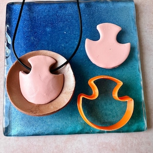 May include: A pink pendant necklace with a black cord, a pink clay pendant, and an orange plastic pendant cutter sit on a blue and green surface. The pendant has a unique, curved shape.