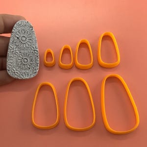 May include: A gray, textured clay stamp with a floral design and several orange teardrop-shaped cookie cutters of varying sizes. The stamp is held by a hand. The cutters are arranged on a peach-colored surface.