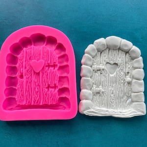 May include: A pink silicone mold and a white casting of a fairy door. The door features a heart-shaped window and a wood-grain texture. The mold is in the shape of the door with a stone archway.