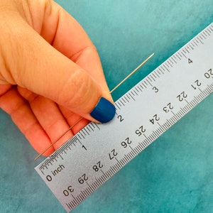 Puede incluir: Primer plano de una regla de plata con marcas negras que mide un alambre delgado de color dorado. La regla est&aacute; marcada en pulgadas y muestra medidas de 0 a 30 pulgadas.