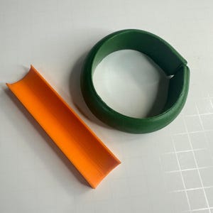 Könnte beinhalten: Zwei 3D-gedruckte Objekte, eines ist ein grüner Ring und das andere ein orangefarbenes Rohr.