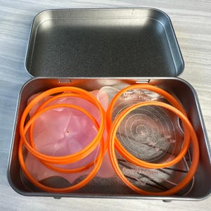以下が含まれることがあります： 金属製の缶に入ったオレンジ色のプラスチック製のキルト定規のセット。定規には「マジックパイ」と表示されています。