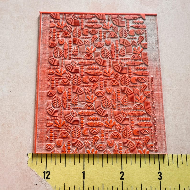 May include: A rectangular, orange rubber stamp with a repeating pattern of stylized botanical shapes. The design includes curved lines, leaf-like forms, and semi-circles. The stamp measures approximately 3 inches wide.