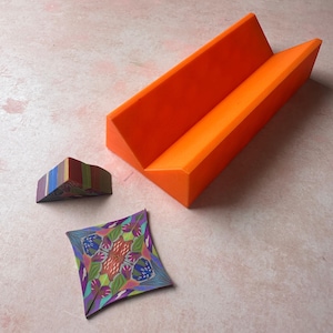 May include: An orange, angular, geometric sculpture with a V-shaped design. Beside it are two smaller, colorful, geometric art pieces. One is a small, multi-colored, curved shape, and the other is a square with a floral pattern.