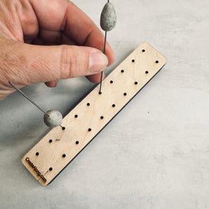 Può includere: Una tavola di perline di legno con più fori, utilizzata per l'artigianato. Due perline grigie macchiate su fili sono inserite nella tavola. Il testo "CreateAlong.com" è stampato sulla tavola.