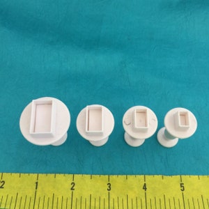 Puede incluir: Conjunto de cuatro cortadores de galletas rectangulares de pl&aacute;stico blanco de diferentes tama&ntilde;os. Los cortadores est&aacute;n sobre una superficie azul con una regla amarilla para la escala.