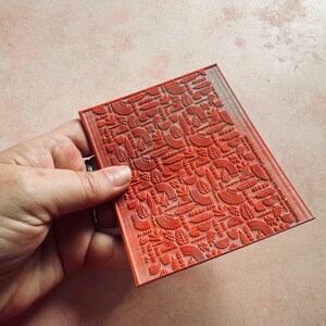 May include: A red rectangular stamp with a detailed, repeating pattern of stylized leaves, arches, and other organic shapes. The stamp is held in a hand, showcasing the intricate design.