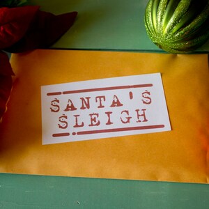May include: A brown envelope with a white label that says "SANTA'S SLEIGH" in red letters. The envelope is on a green surface with red and green ornaments.