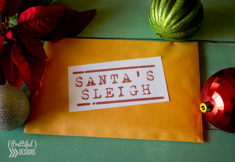May include: A brown envelope with a white label that says "SANTA'S SLEIGH" in red letters. The envelope is on a green surface with red and green ornaments.