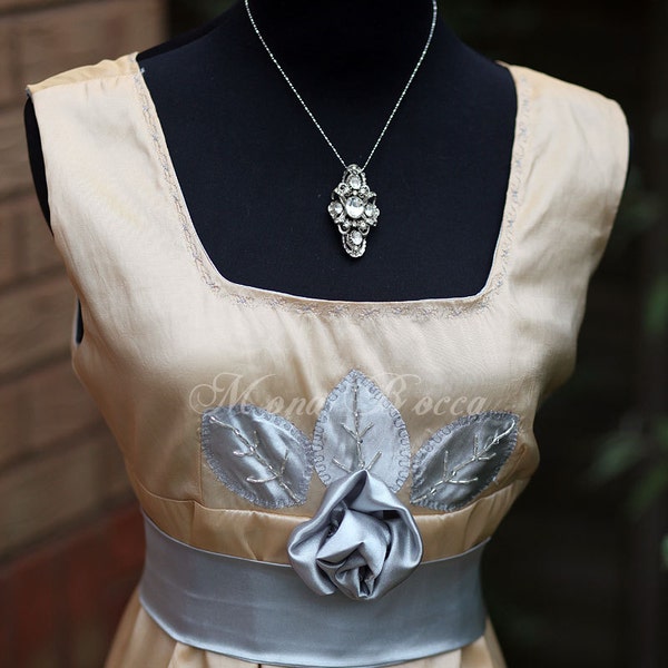 Puede incluir: Un vestido sin mangas de color beige claro con una faja de satén plateado y un diseño floral bordado en plata. El vestido tiene un escote cuadrado y un collar con un colgante de cristal.