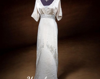 May include: A cream-coloured, floor-length dress with short sleeves and a V-neck. The dress features intricate silver embroidery on the bodice, waist, and skirt. A delicate silver necklace adorns the mannequin's neck. The website address Monabocca.com is visible.