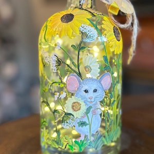 以下が含まれることがあります： ひまわりとデイジーの花畑の中にネズミが描かれたガラス瓶。瓶には蝶が飾られており、中には電球の紐が入っています。