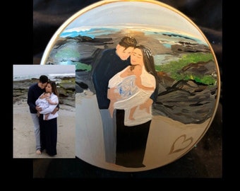 May include: A hand-painted Christmas ornament featuring a family of three standing on a beach. The ornament is white with a gold trim and a heart shape on the bottom. The family is painted in a realistic style and the background is a blue and green seascape.