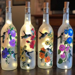 May include: Four frosted glass bottles with cork tops, each painted with a hummingbird and flowers. The bottles are lit from within with white fairy lights.