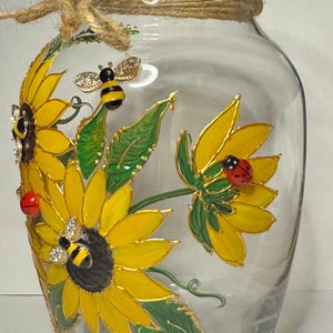 Puede incluir: Jarrón de cristal transparente decorado con girasoles amarillos, hojas verdes y pequeñas mariquitas y abejas. El borde está envuelto con cuerda. Los girasoles tienen contornos dorados y las abejas tienen detalles de pedrería. Un jarrón floral decorativo.