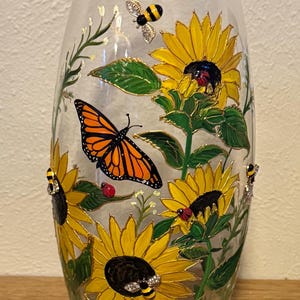 May include: A clear glass vase with a hand-painted design featuring yellow sunflowers, green leaves, and a monarch butterfly. Decorative bees and ladybirds are also included in the design. The vase has a gold trim.