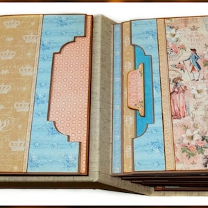 May include: A handmade scrapbook with a vintage theme. The pages feature illustrations of people in period clothing, crowns, and floral patterns. The color palette includes shades of blue, peach, and tan, with brown borders.
