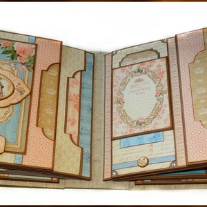 May include: Open scrapbook with a vintage design. The pages feature a mix of light blue, pink, and beige backgrounds with floral and crown motifs. Decorative labels and frames are present, with a central oval image.