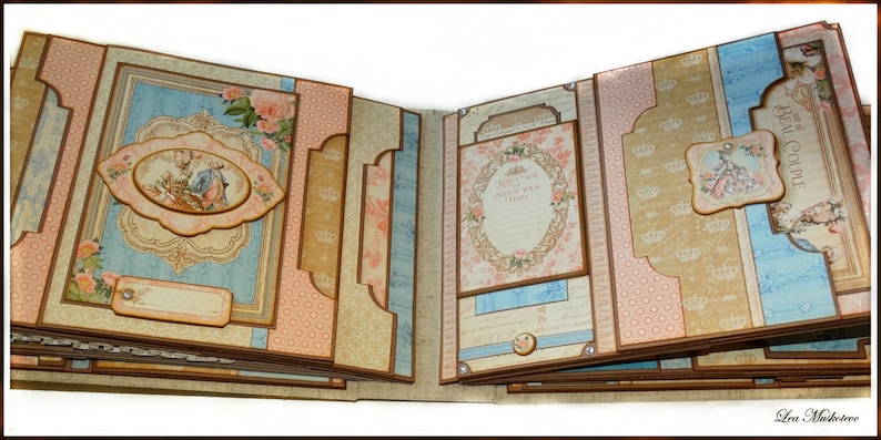 May include: Open scrapbook with a vintage design. The pages feature a mix of light blue, pink, and beige backgrounds with floral and crown motifs. Decorative labels and frames are present, with a central oval image.