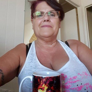 Pu&ograve; includere: Una donna con gli occhiali e una canottiera bianca tiene in mano una tazza di ceramica bianca con un'immagine colorata di un viso maschile. La tazza ha un manico nero. La donna sorride.