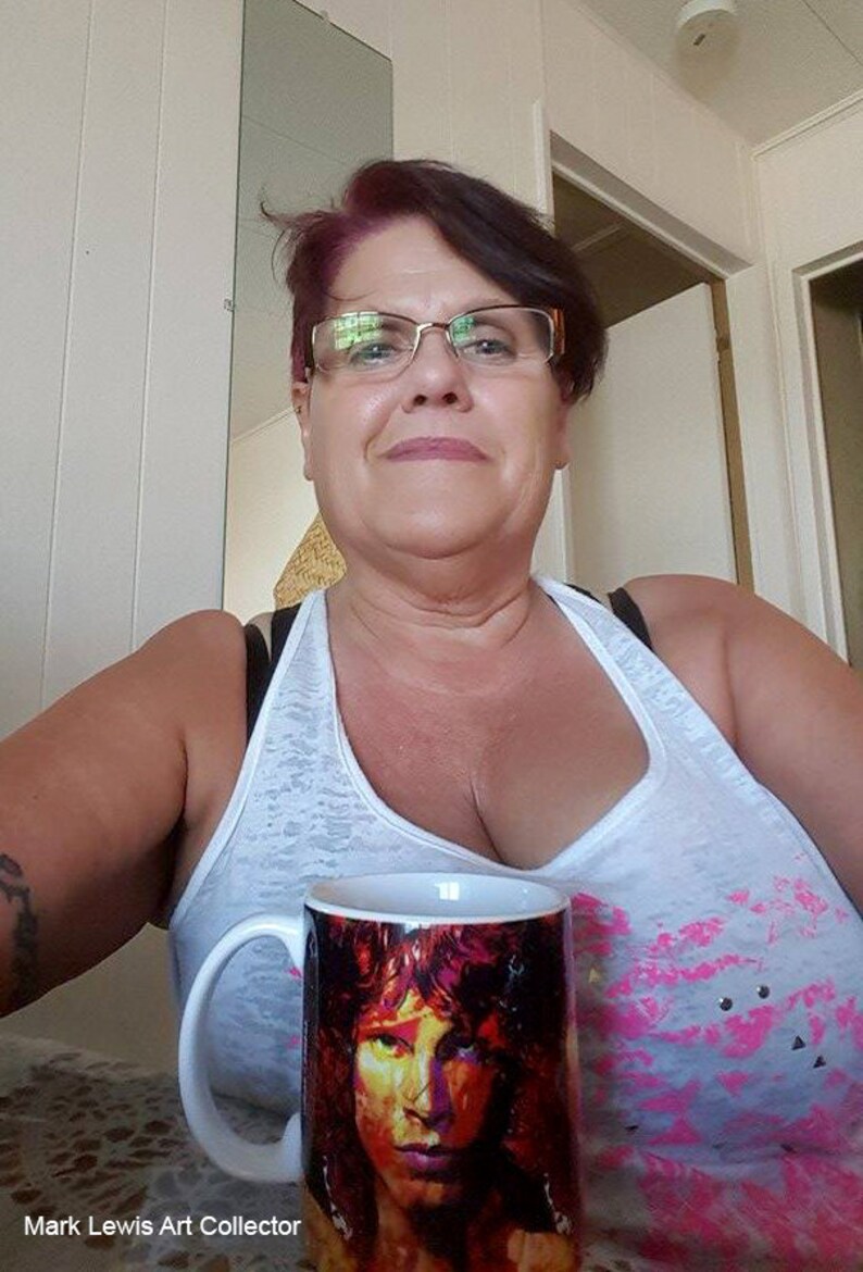 Pu&ograve; includere: Una donna con gli occhiali e una canottiera bianca tiene in mano una tazza di ceramica bianca con un'immagine colorata di un viso maschile. La tazza ha un manico nero. La donna sorride.