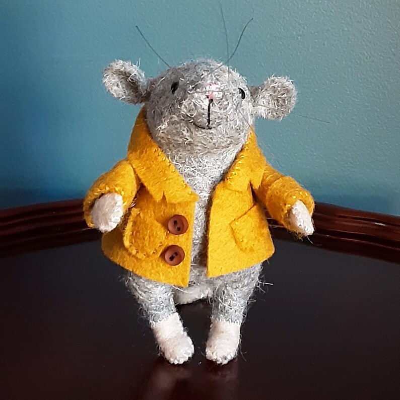 May include: A gray felt mouse wearing a yellow felt coat with brown buttons. The mouse has a pink nose and white feet.