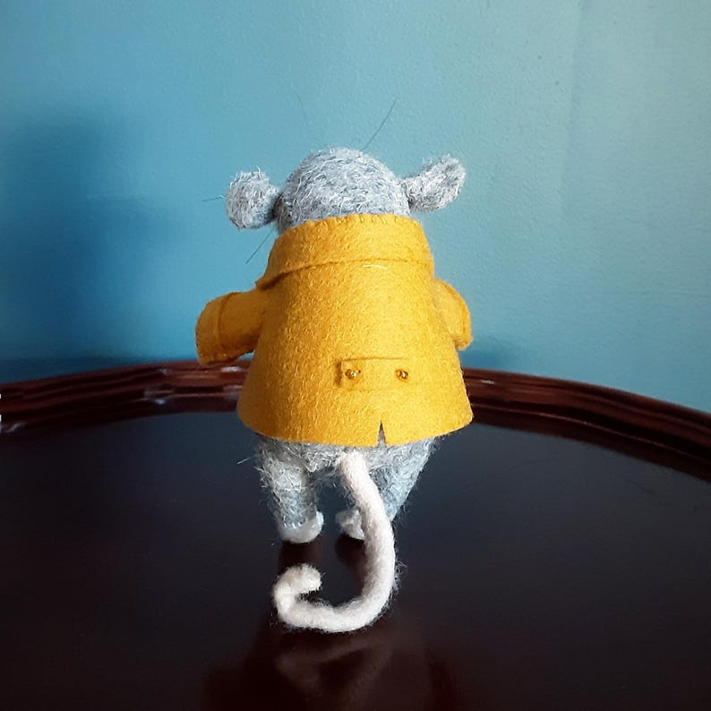May include: A gray felt mouse wearing a yellow felt coat with two buttons on the back. The mouse has a white felt tail.