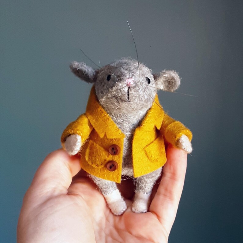 May include: A small gray felt mouse wearing a yellow felt coat with brown buttons. The mouse is sitting in a hand.