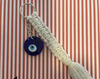 Puede incluir: Llavero de macramé blanco con un amuleto de ojo turco azul. El llavero tiene un borla al final. El fondo es de rayas rojas y blancas. El texto "CRAFTYDEVIL CRAFTS" está en la parte inferior de la imagen.