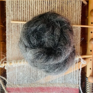 May include: A close-up of a wooden weaving loom with a ball of dark gray yarn. The yarn is fluffy and textured. The loom has vertical and horizontal threads, with a partially woven section in the foreground.