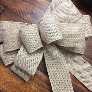 May include: A large, decorative bow made of tan burlap fabric. The bow is centered in the frame, with multiple loops and long, flowing tails. The coarse texture of the burlap is visible, and the bow is set against a wooden surface.
