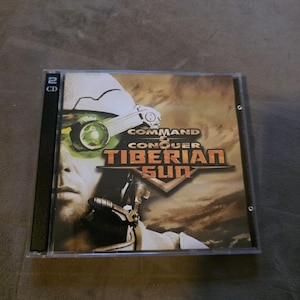 May include: A Command & Conquer: Tiberian Sun PC game in a clear plastic case. The cover art features a futuristic soldier with a green-tinted visor and the game's title in bold, metallic lettering. The case indicates it contains two CDs.