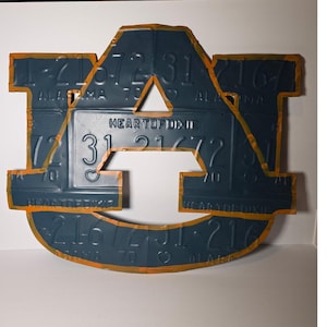 May include: A dark blue metal Auburn University logo, crafted from license plates, with orange painted edges. The logo features the text "HEART OF DIXIE" and various numbers and letters from the license plates. A unique piece of collegiate decor.