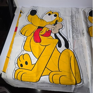 May include: A fabric panel featuring a yellow Pluto cartoon character with a red tongue and a bone. The character is outlined in black on a white background. Instructions are printed on the fabric, including "LEAVE OPEN FOR STUFFING".