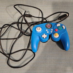 May include: A blue video game controller with a long black cord. The controller features a white directional pad, a yellow analog stick, a green analog stick, a red button, two white buttons, and a central emblem featuring a red and blue shield.