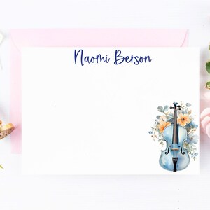 May include: A white stationery card with the name "Naomi Berson" printed in blue ink. The card features a watercolor illustration of a blue violin with floral accents.