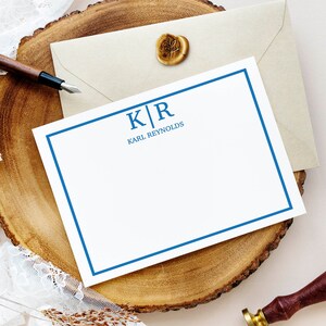 Könnte beinhalten: Eine weiße Karte mit einem blauen Rand und den Initialen "K|R" in Blau, gefolgt vom Text "KARL REYNOLDS". Die Karte liegt auf einer Holzoberfläche mit einem braunen Umschlag und einem Wachssiegel.