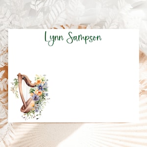 May include: A white card with the name "Lynn Sampson" printed in green cursive script. The card features a watercolour illustration of a wooden harp with yellow, orange, and purple flowers.