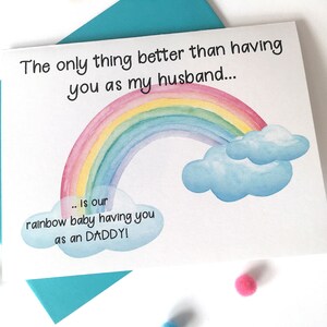 May include: A white card with a rainbow and clouds illustration. The text on the card reads "The only thing better than having you as my husband... is our rainbow baby having you as an DADDY!"