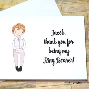 May include: A white card with a cartoon illustration of a boy in a suit holding a pillow. The text on the card reads "Jacob, thank you for being my Ring Bearer!"
