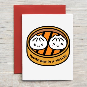 May include: A white card with a cartoon illustration of two steamed buns in a bamboo steamer. The text "You're bun in a million" is written below the steamer.
