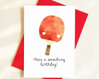 Op de afbeelding: Een witte wenskaart met een illustratie van een rode pingpongbat met een lachend gezicht en de tekst "Have a smashing birthday!". De kaart ligt op een rode envelop. De bat heeft een bruin handvat.